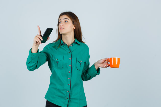 皮肤年轻的女士拿着桔黄色的茶杯和手机 指着衬衫 小心地看着前视图杯子感性头发