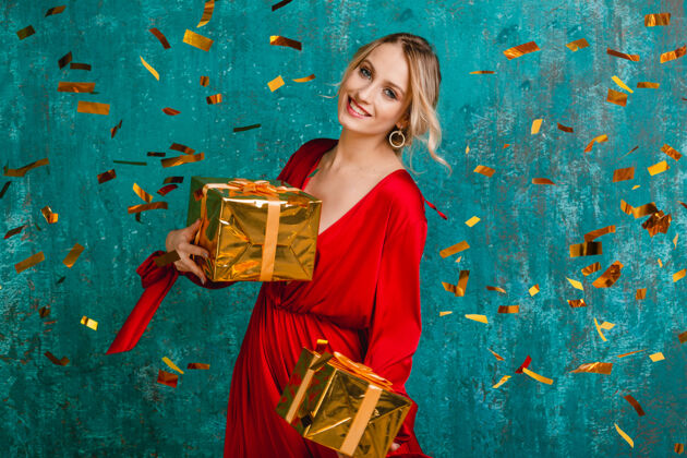 年迷人的快乐微笑的女人穿着时尚的红色连衣裙 用礼物庆祝圣诞节和新年五彩纸屑举行乐趣