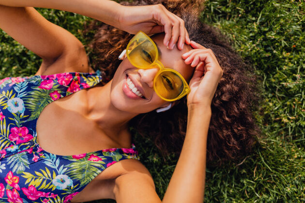 混血儿年轻时尚的黑人女子 戴着无线耳机听音乐 躺在公园的草地上玩得很开心 夏日的时尚风格 五颜六色的时髦装扮 俯瞰风景年轻耳机黄色