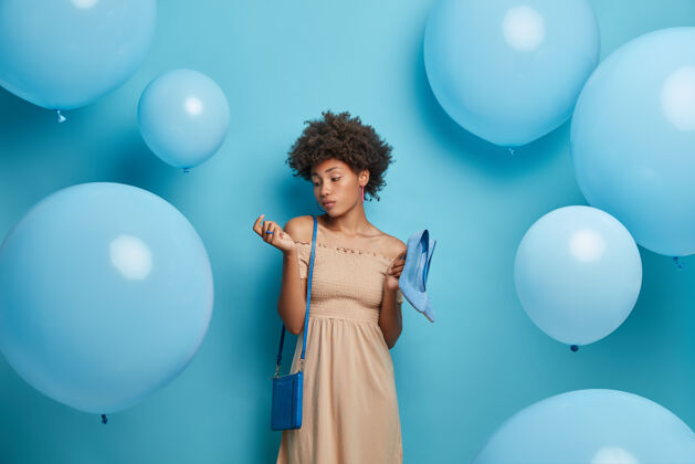 服装严肃时尚的女人看着她新修的美甲 穿着米色裙子 带着蓝色高跟鞋 适合特殊场合的包裙 周围环绕着蓝色气球女性特别鞋