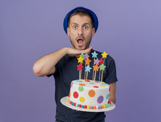 兴奋戴着蓝色帽子的白人帅哥兴奋地把手放在下巴下面 手里拿着生日蛋糕 背景是紫色的 上面留有复制空间男人手空间