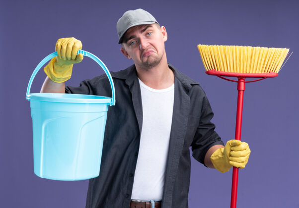 拖把悲伤的歪头年轻帅气的清洁工穿着t恤和帽子 戴着玻璃 拿着桶和拖把孤立在紫色的墙上举行悲伤桶
