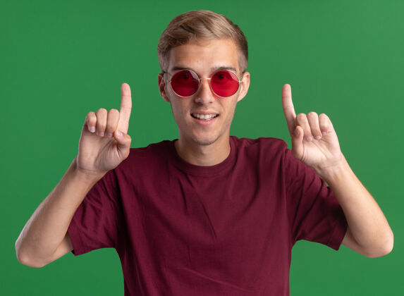 微笑面带微笑的年轻帅哥穿着红衬衫 戴着眼镜 指着绿色的墙上孤立起来穿年轻点