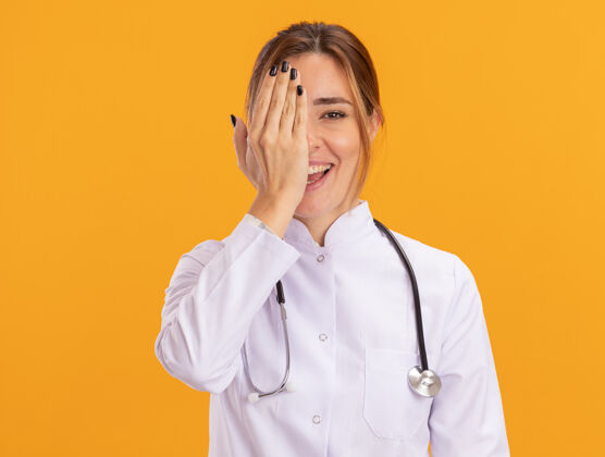 罩子微笑的年轻女医生穿着医用长袍 听诊器遮住眼睛 手被隔离在黄色的墙上听诊器医生医疗