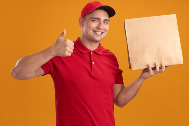 微笑笑容满面的年轻送货员穿着制服 戴着帽子 拿着纸食品包 竖起大拇指 孤立地站在橙色的墙上送货抱着纸张
