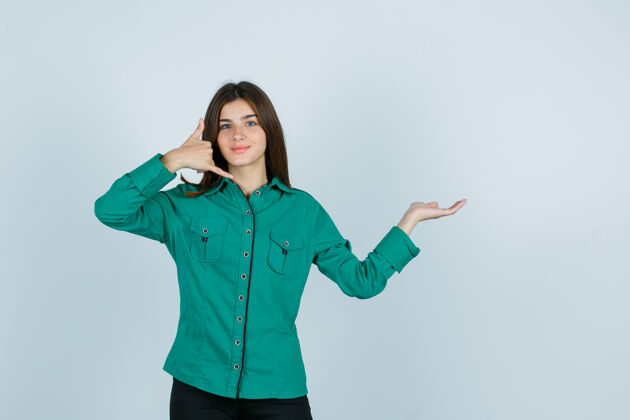 年轻一个穿着绿色上衣 黑色裤子的年轻女孩 展示着打电话的姿势 把手掌放在一边 看上去很乐观 正对着前方表演自然欢呼