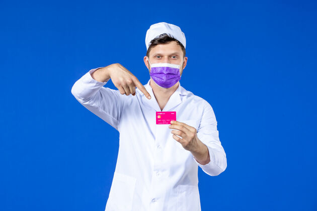 信用前视图中的男医生穿着医疗服和面具持有信用卡的蓝色病毒药品男医生
