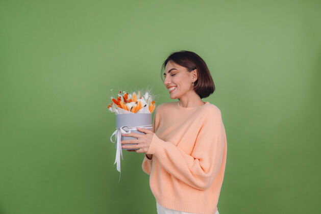 时尚年轻女子穿着休闲桃色毛衣隔离在绿橄榄墙上手持橙白色花盒组成的棉花 吉普赛拉小麦和拉古鲁斯作为礼物开心惊喜美丽欢乐年轻