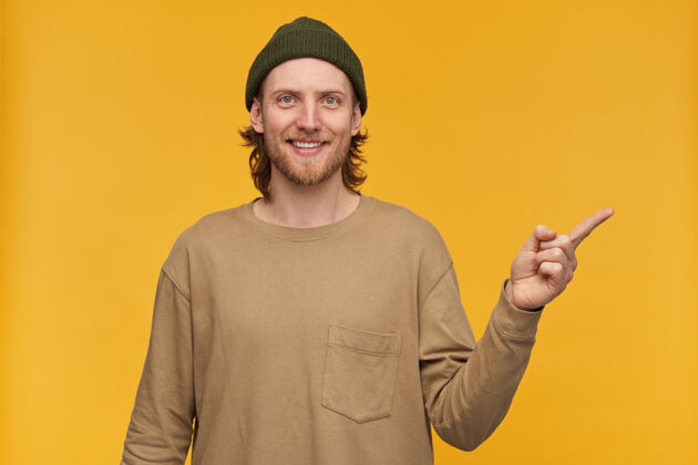 男人一个乐观的男人 金发 胡子 小胡子 穿着绿色的小豆豆和米色的毛衣手指指向右边的复制空间 隔离在黄色的墙上情绪休闲毛衣