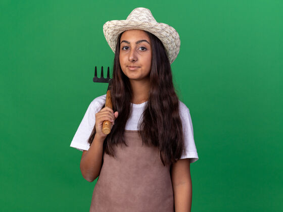 年轻快乐的年轻园丁女孩围裙和夏季帽子举行迷你耙脸上带着微笑站在绿色的墙壁