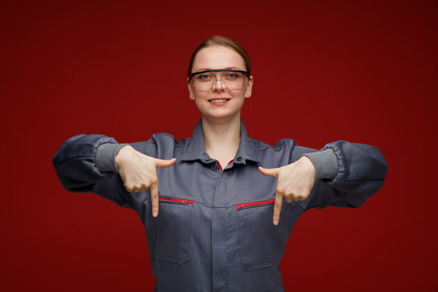 安全微笑着的年轻金发女工程师穿着制服 戴着安全眼镜指着下面年轻制服指尖