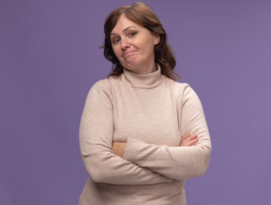年龄身穿米色套头衫 双臂交叉 表情怀疑的中年妇女站在紫色的墙上表情高领毛衣中年