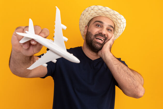 帽子胡子男人穿着黑色t恤 戴着夏天的帽子 手里拿着玩具飞机 站在橘色的墙上 惊奇而快乐地微笑着欢呼惊奇男人