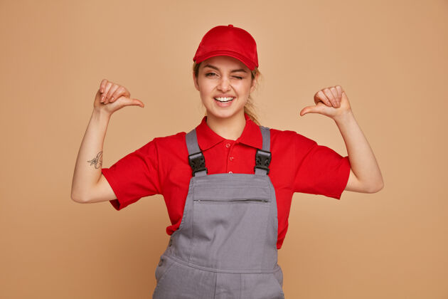 制服快乐的年轻女建筑工人穿着制服 戴着帽子指着自己眨眼年轻工人喜悦