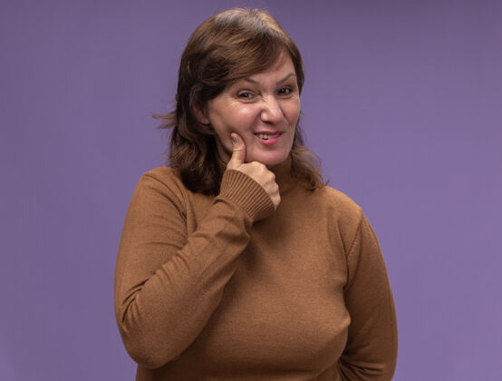 年龄穿着棕色高领毛衣的中年妇女站在紫色的墙上 摸着她的脸颊 脸上挂着牙痛中年摸站着