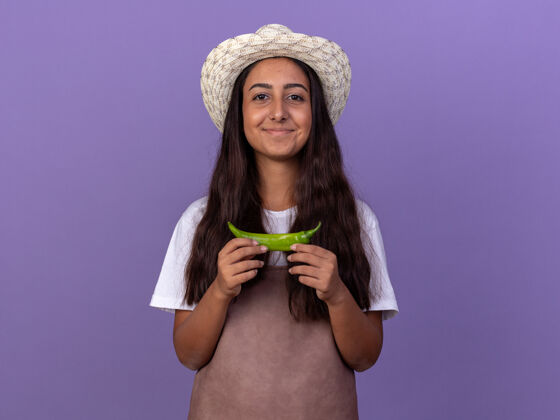 帽子穿着围裙 戴着夏帽 手拿青椒的年轻园丁女孩站在紫色的墙上 脸上洋溢着幸福的笑容围裙站辣椒