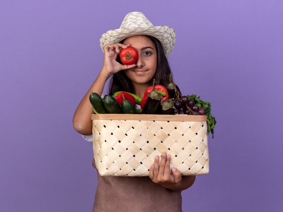 板条箱年轻的园丁女孩围着围裙 戴着夏帽 手里拿着装满蔬菜的板条箱 站在紫色的墙上 用新鲜的西红柿遮住眼睛花园全套新鲜