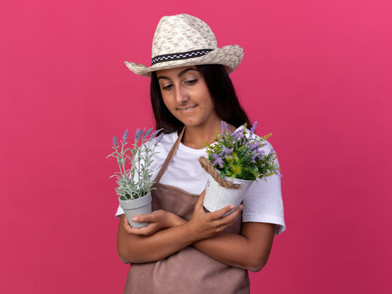 锅年轻的园丁女孩围着围裙 戴着夏帽 手里拿着盆栽植物 站在粉红色的墙上 脸上带着微笑 看上去很自信帽子微笑花园