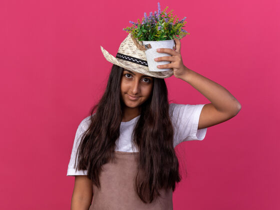 围裙穿着围裙 戴着夏帽的年轻园丁女孩 头上抱着盆栽植物 脸上带着微笑站在粉色的墙上年轻花园帽子