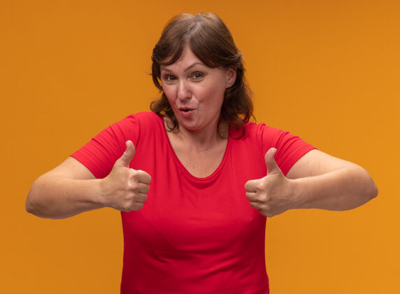 中间身着红色t恤的中年妇女站在橙色的墙上微笑着竖起大拇指拇指站立女人