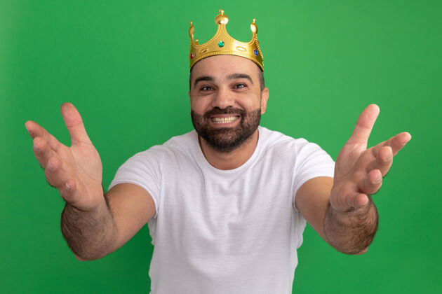 男人满脸胡须 身穿白色t恤 头戴金色皇冠的男子站在绿色的墙边 高兴而积极地伸出双手 做着欢迎的手势制造积极立场