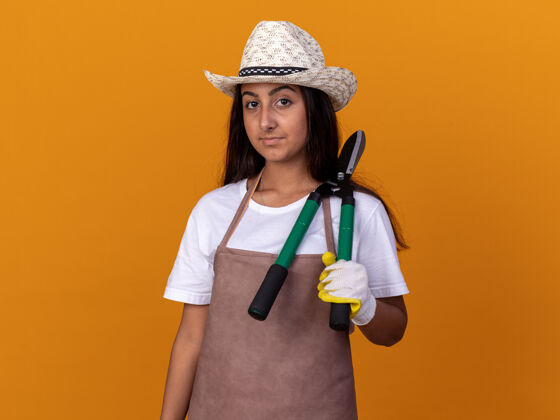 严肃穿着围裙 戴着夏帽 手持树篱修剪器 表情严肃的年轻园丁女孩站在橙色的墙上帽子剪子围裙