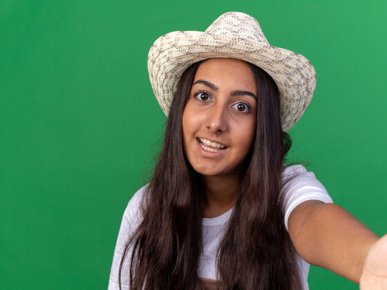站着戴着围裙和夏帽的快乐的年轻园丁女孩站在绿色的墙上 快乐而欢快地微笑着年轻的帽子围裙