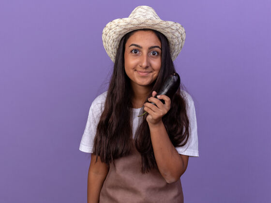 年轻的穿着围裙 戴着夏帽 抱着茄子的年轻园丁女孩站在紫色的墙上 快乐而积极地微笑着围裙积极的茄子