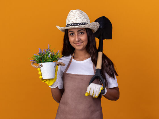 铲子年轻的园丁女孩拿着盆栽和铲子 脸上带着微笑站在橙色的墙上微笑年轻人花园