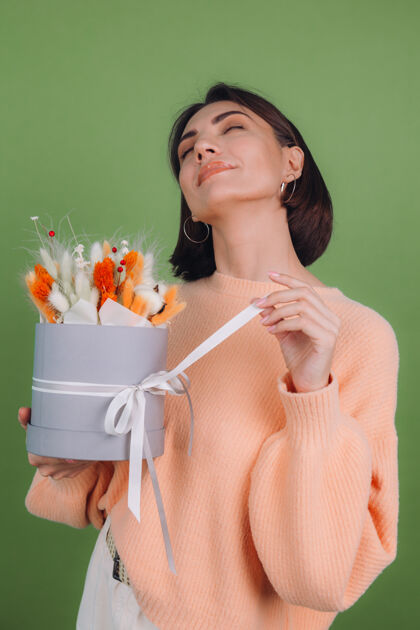 礼盒年轻女子穿着休闲桃色毛衣隔离在绿橄榄墙上手持橙白色花盒组成的棉花 吉普赛拉小麦和拉古鲁斯作为礼物开心惊喜女人花圆形盒