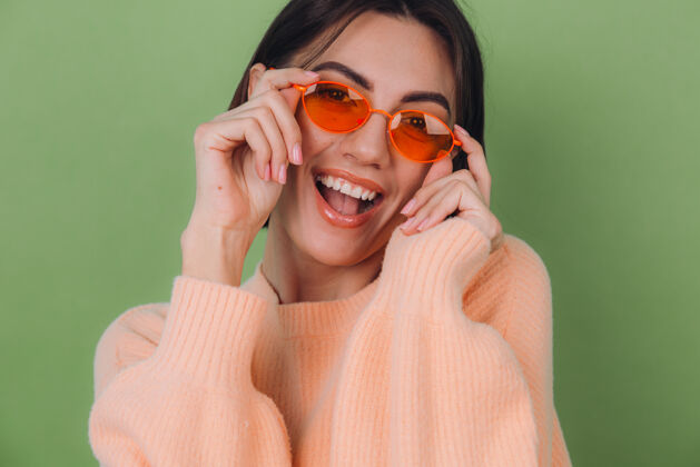 女人年轻时尚的女士穿着休闲的桃色毛衣 戴着橙色的眼镜 隔着绿橄榄色的墙壁 开心的 积极的笑着 围着复制空间转脸快乐黑发