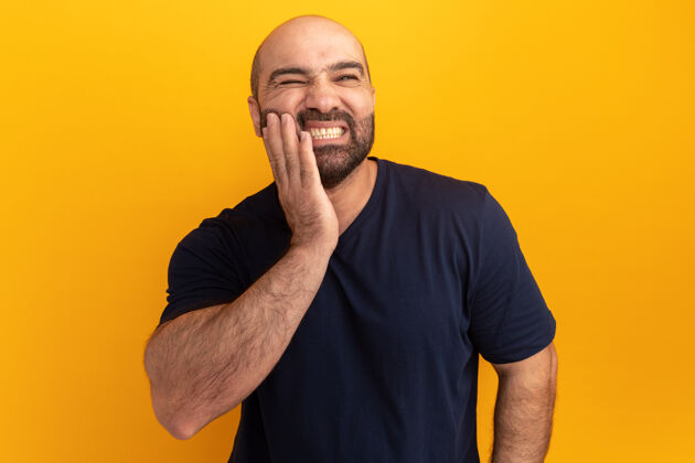 表情一个留着大胡子的男人穿着海军蓝t恤 站在橘色的墙上 看着不舒服 摸着他的脸颊 牙疼胡须触摸不舒服