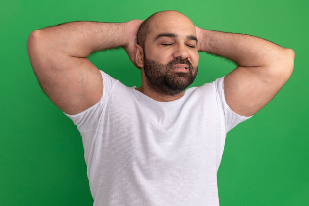 闭上一个留着胡子的男人 穿着白色t恤 闭着眼睛 双手放在脑后 自信地站在绿色的墙上表情站着男人