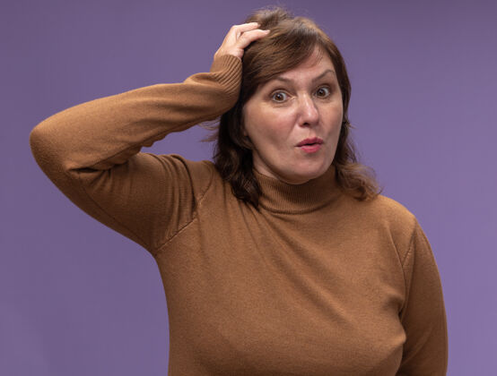 错误穿棕色高领套头衫的中年妇女误站在紫色的墙上 把手放在头上弄糊涂了年龄困惑头