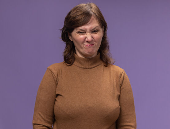 品牌穿着棕色高领毛衣的中年妇女站在紫色的墙上做鬼脸 既不高兴又不高兴中等鬼脸不开心
