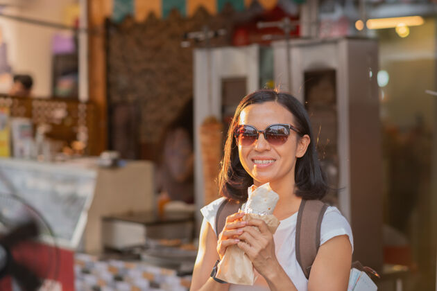 街头食品快乐的女人在街上吃着带馅儿的烤肉串市场背包城市