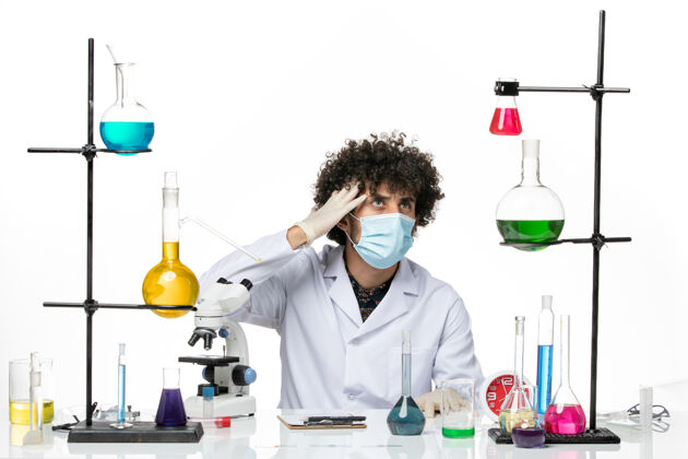 男性化学家正面图：男药剂师穿着医疗服 戴着口罩 坐在白色的桌子上 拿着不同的溶液医学面罩坐着