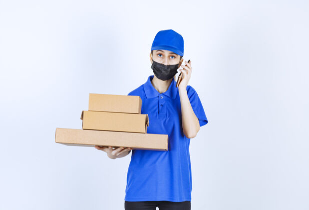 年轻戴着面具 穿着蓝色制服的女快递员手里拿着一堆纸箱 对着电话说话智能娱乐成人