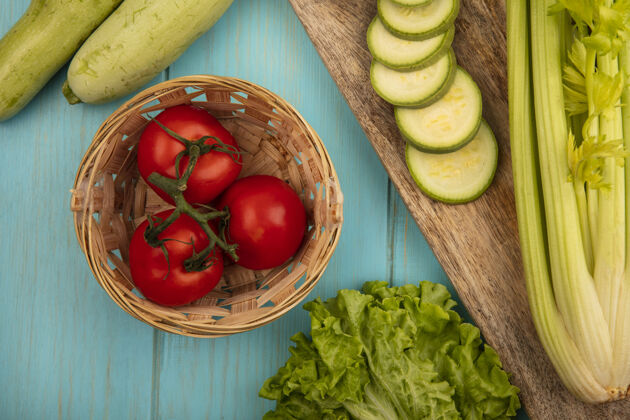 木材一桶新鲜西红柿的顶视图 切碎的西葫芦放在木制的厨房板上 芹菜和莴苣被隔离在蓝色的木制表面上饮食蔬菜板