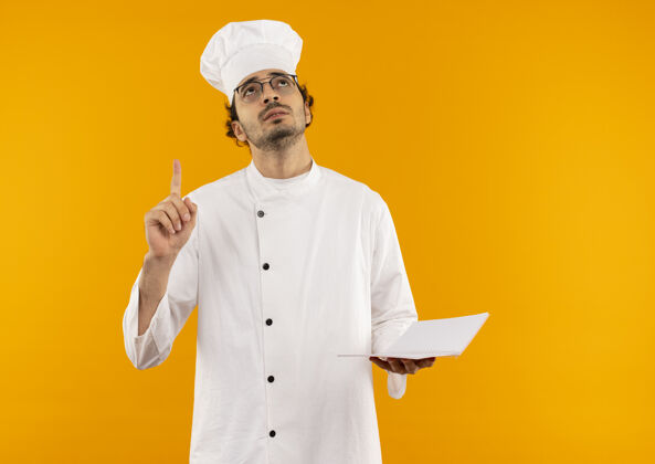 向上看着抬头思考的年轻男厨师穿着厨师制服 戴着眼镜拿着笔记本 指着上面思考烹饪年轻