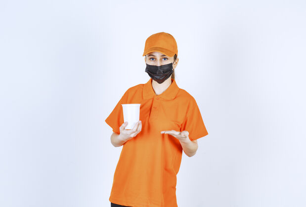 工人穿着黄色制服 戴着黑色面具的女快递员拿着一个塑料外卖杯指着它年轻质量模特