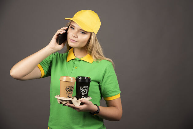 人年轻的女送货员拿着几杯咖啡在智能手机上讲话手机模特女孩