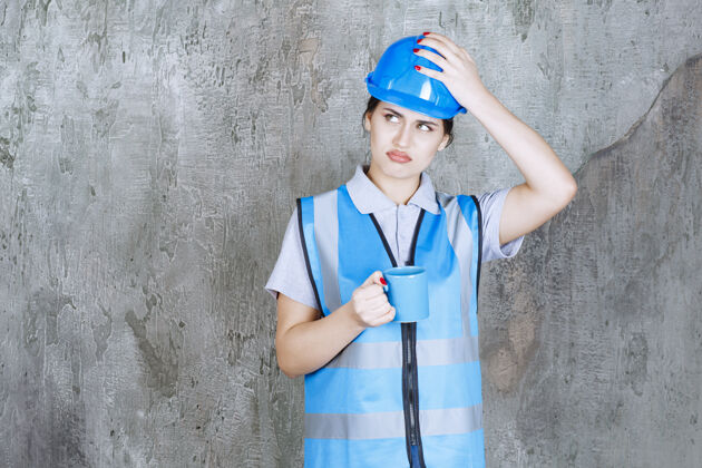 消极身穿蓝色制服 头戴安全帽 手持蓝色茶杯的女工程师 看上去既困惑又紧张模特人体模特工人