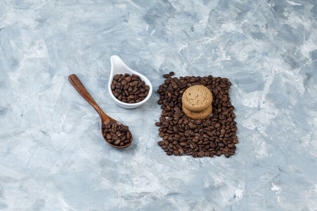 拿铁高角度看咖啡豆在木制勺子 白色陶瓷壶饼干浅蓝色大理石背景水平早晨木头角度