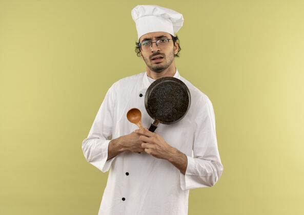 眼镜悲伤的年轻男厨师穿着厨师制服 戴着眼镜 拿着勺子拿着平底锅厨师男性绿色