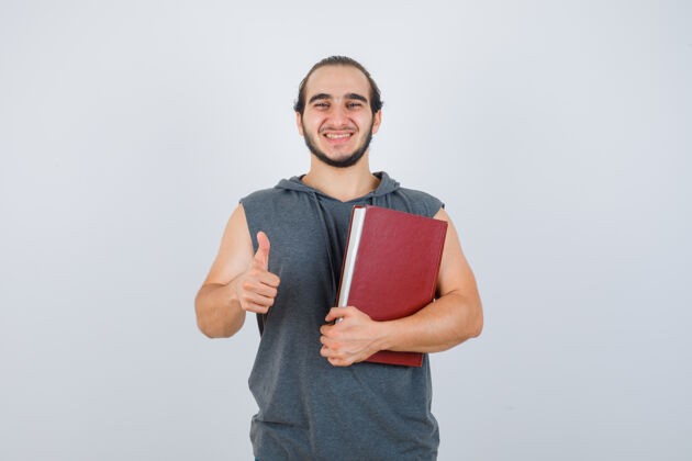 时尚年轻的男性拿着书 穿着无袖连帽衫竖起大拇指 看上去很开心 正面照站立成人男性