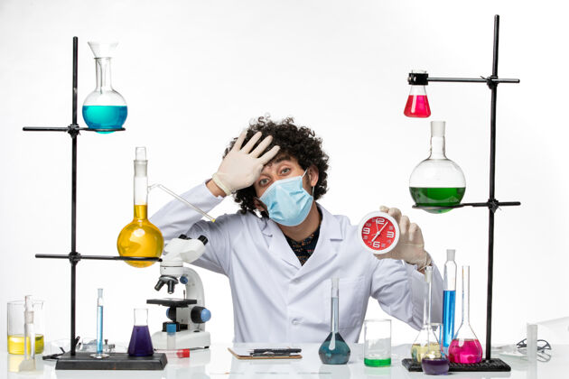 医学正面图：穿着医疗服 戴着面具的男性化学家 在白色的桌子上拿着红色的时钟实验室外套科学