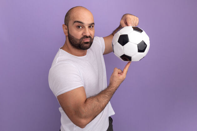 球一个留着胡子 穿着白色t恤的男人 手指上拿着足球 脸严肃地站在紫色的墙上足球手指严肃