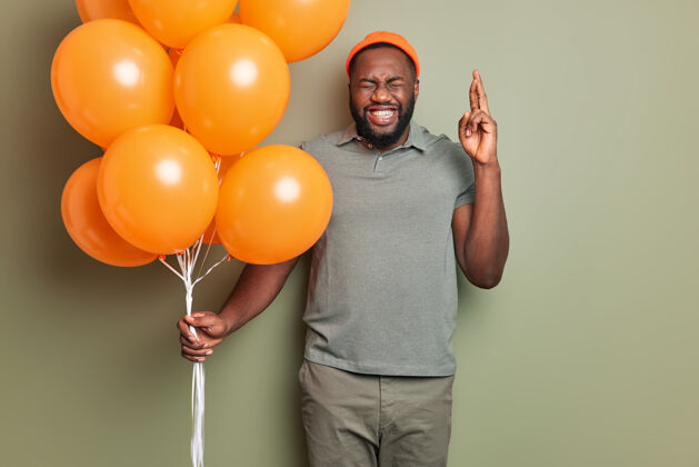 学生满脸胡须的快乐男人在生日的时候用手指交叉许愿 手里拿着一束亮橙色的充气气球 穿着时髦的衣服站在室内男人生日快乐