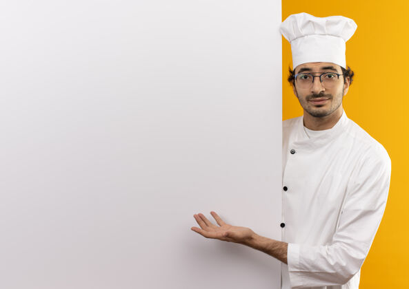 男性高兴的年轻男厨师穿着厨师制服 戴着眼镜 手拿着白色的墙壁 在黄色的墙壁上展示着隔离的复制空间手年轻请